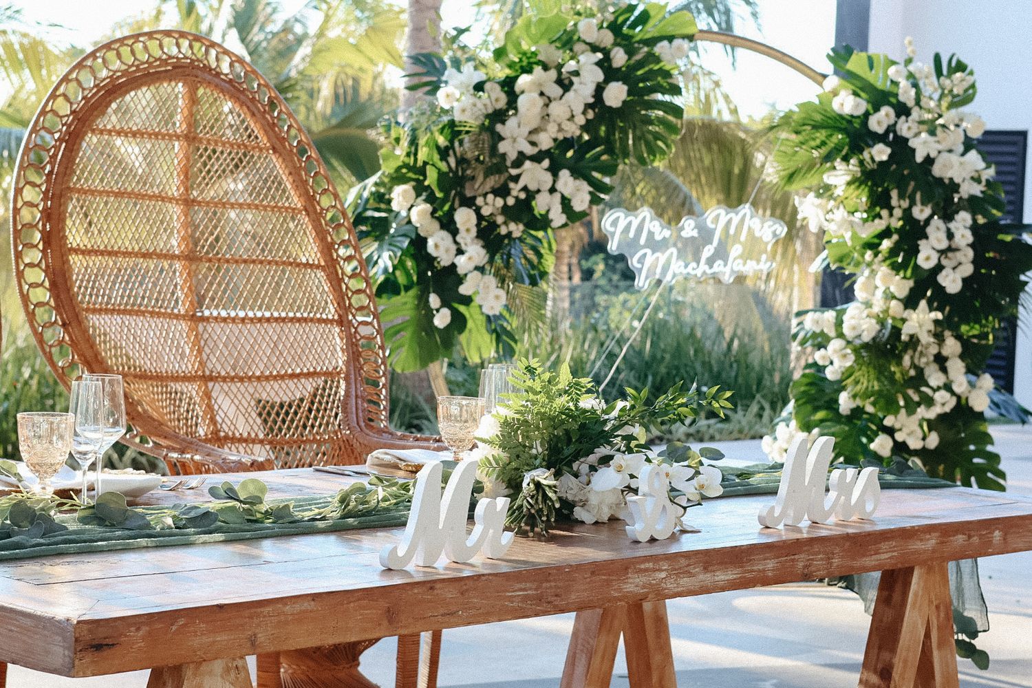 An elegant outdoor wedding reception setup in Mexico, featuring a rustic wooden table, bohemian peacock chairs, and lush white floral arrang
