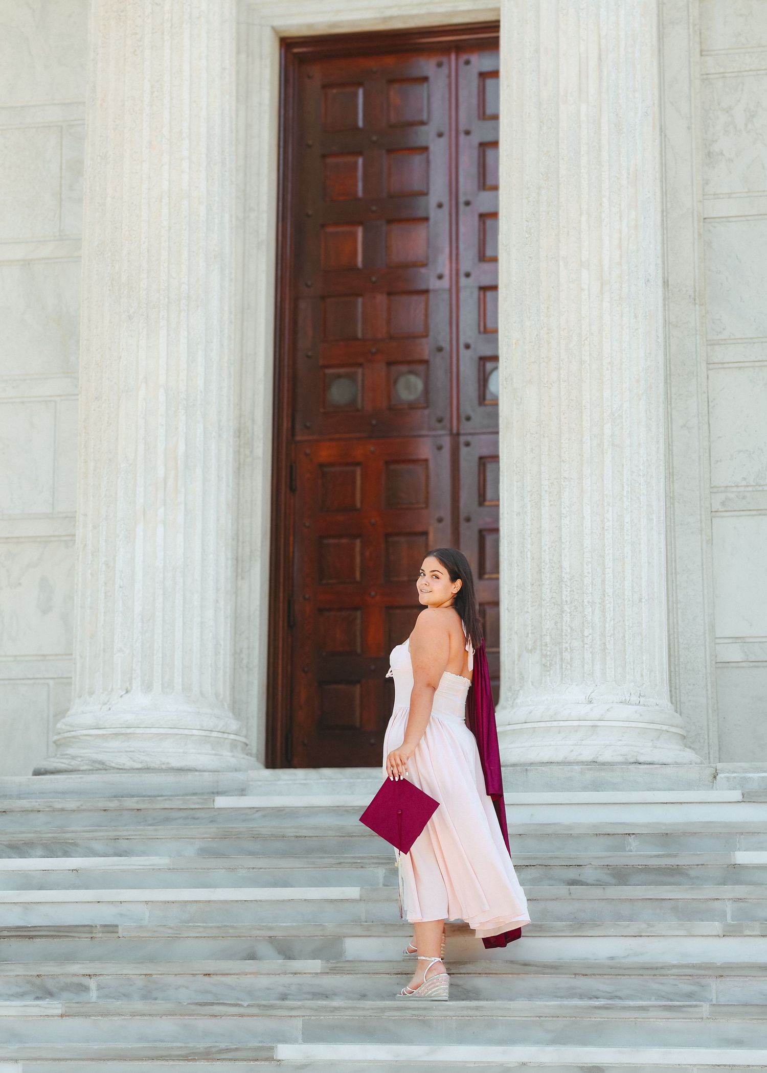 A high school graduate in a pink dress smiling and holding a graduation pennant while posing next to a classic stone column in Princeton