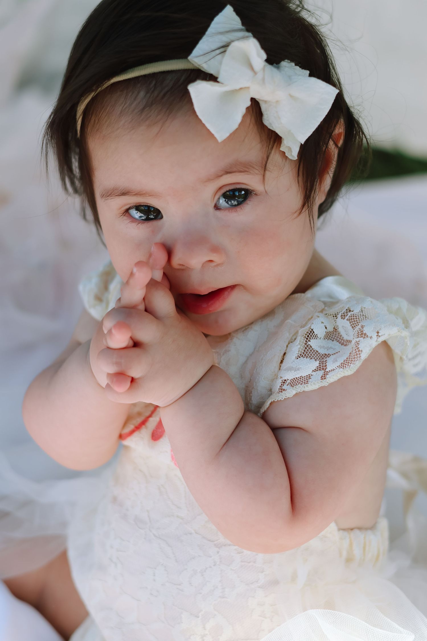A baby girl celebrating her first birthday, wearing a white tulle dress and sitting on a blanket outdoors in a sunlit grassy field.