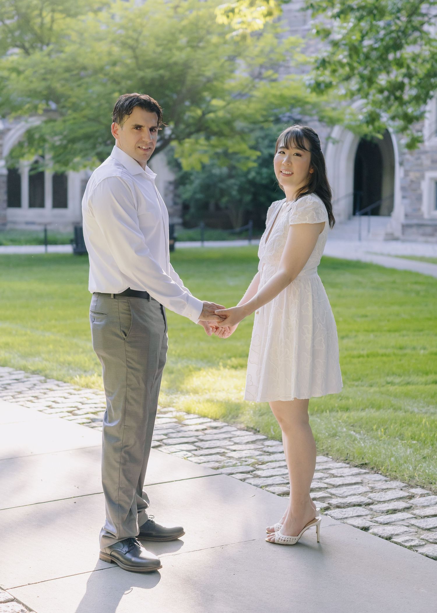 An engaged couple holding hands and walking along a stone path in front of a historic gothic-style building at Princeton University.