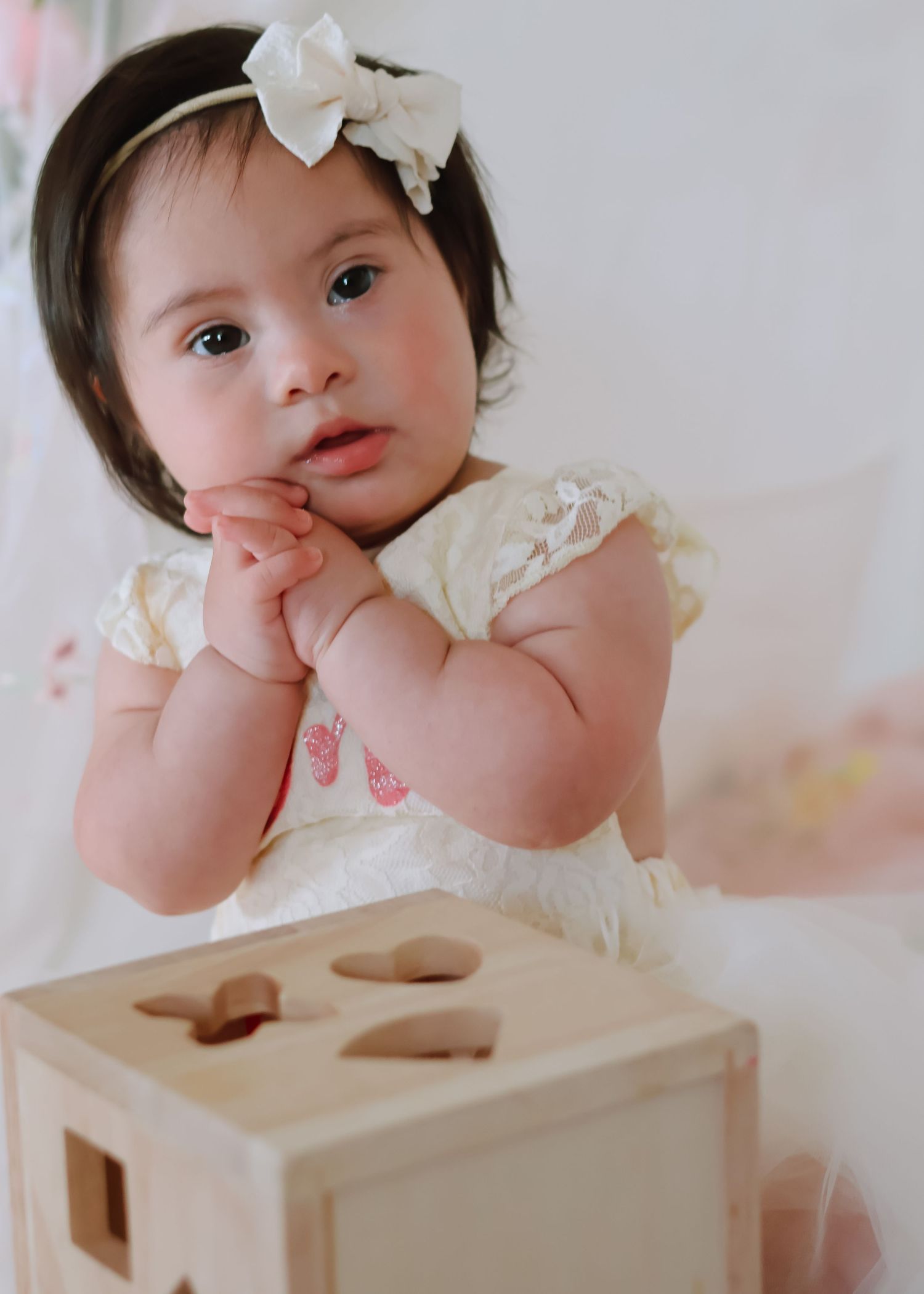 A baby girl celebrating her first birthday, wearing a white tulle dress and sitting on a blanket outdoors in a studio indoors.
