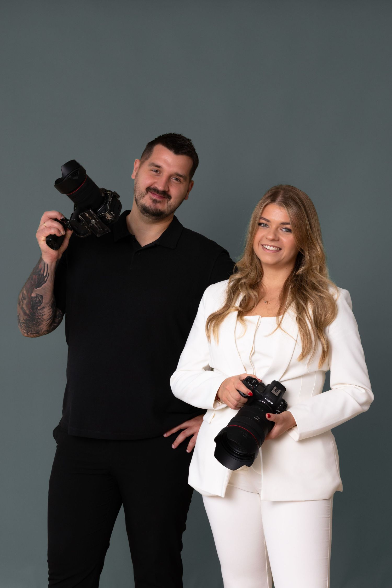 Two professional photographers, man in black and woman in white suit, posing with DSLR cameras against gray background.