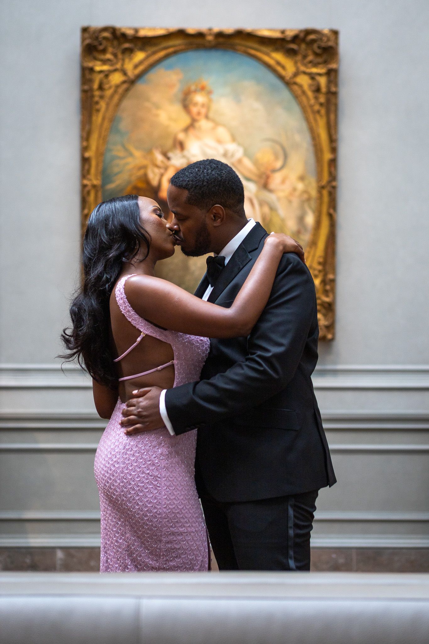 a couple embracing and kissing indoors, with a large, ornate painting visible in the background. The woman wearing a pink dress, engagement.