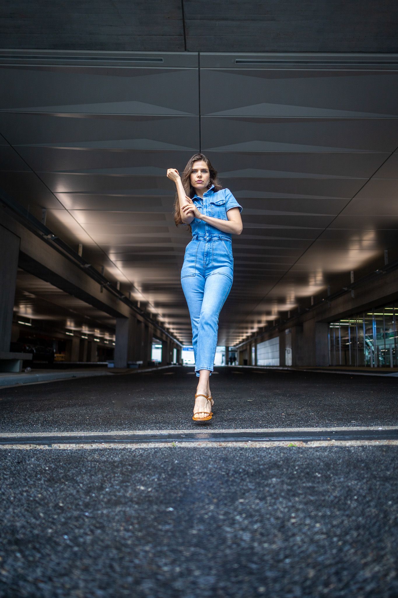 A woman in a blue denim jumpsuit is standing in the middle of a street, framed by modern architecture and leading lines.