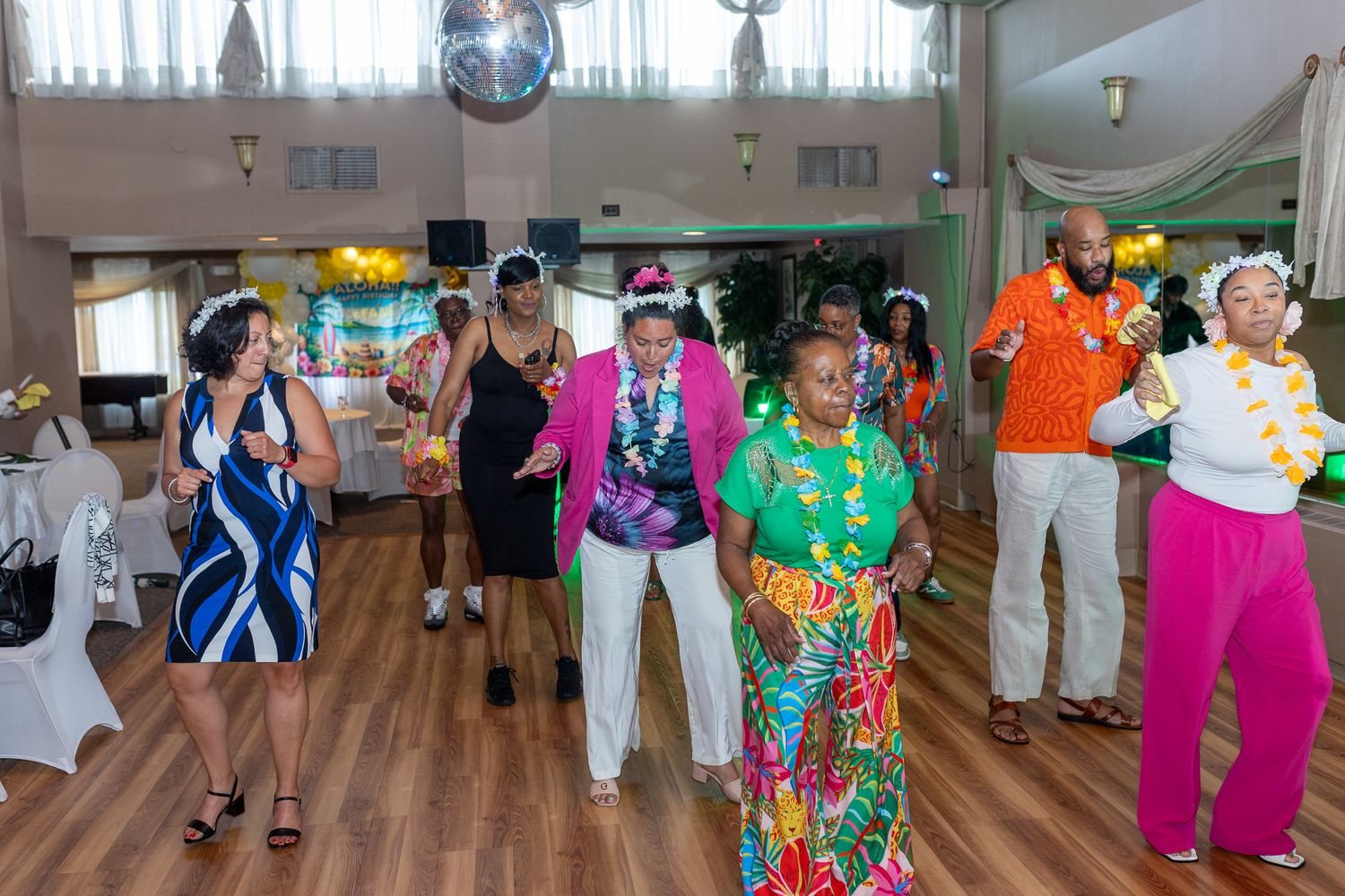 a group of people dancing indoors, likely at a party or celebration. They are wearing colorful, festive clothing, including Hawaiian leis.