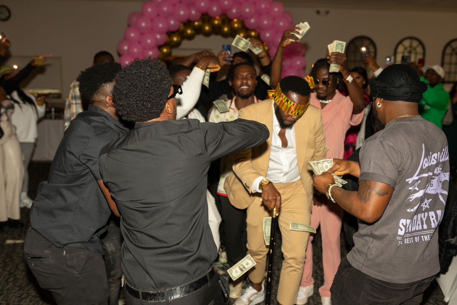 This photo features a group of people celebrating indoors, with a man in a light-colored suit being showered with dollar bills.