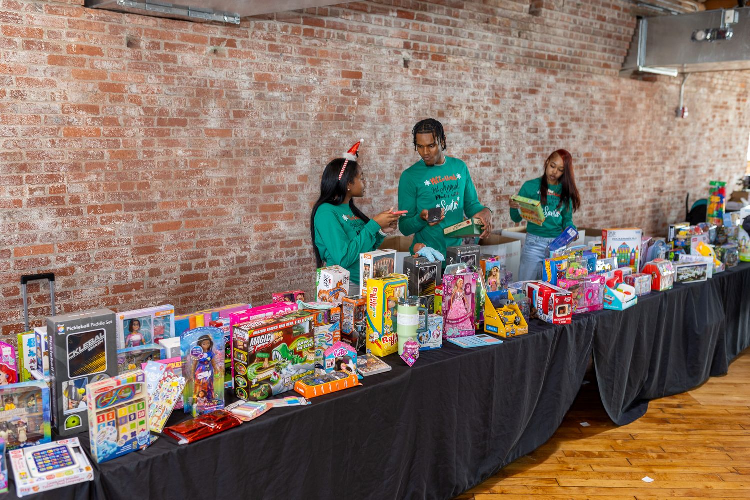 This photo features two women standing behind a long table covered in various wrapped gifts and toys, with a brick wall in the background.