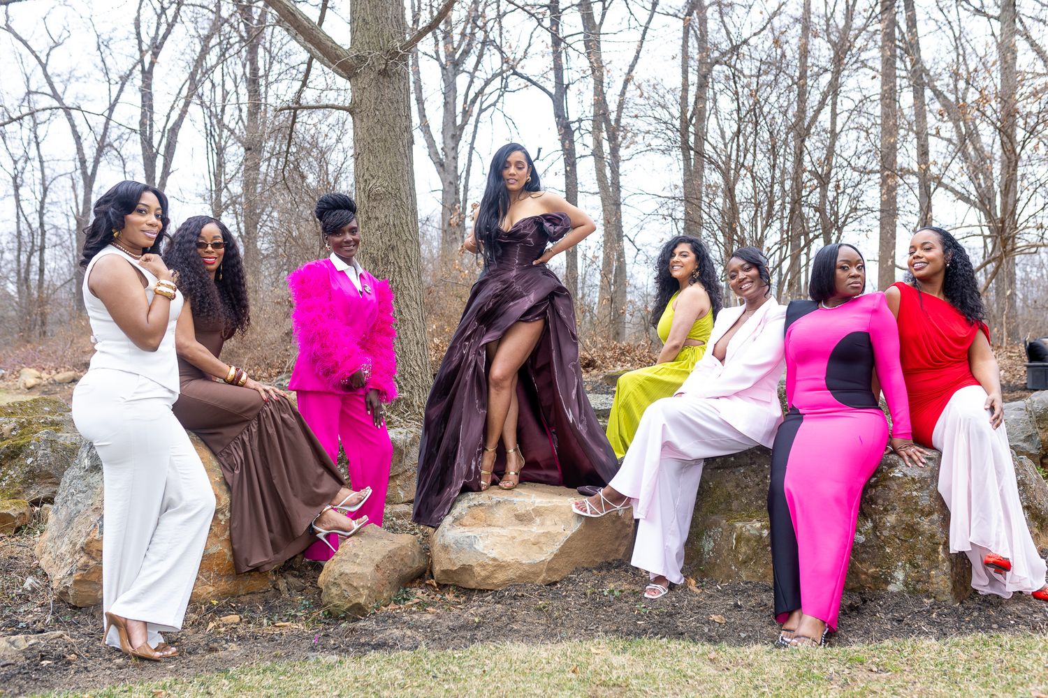 a group of seven women posing outdoors on a rocky terrain with bare trees in the background. Wearing dress of multiple colors, pink, red.
