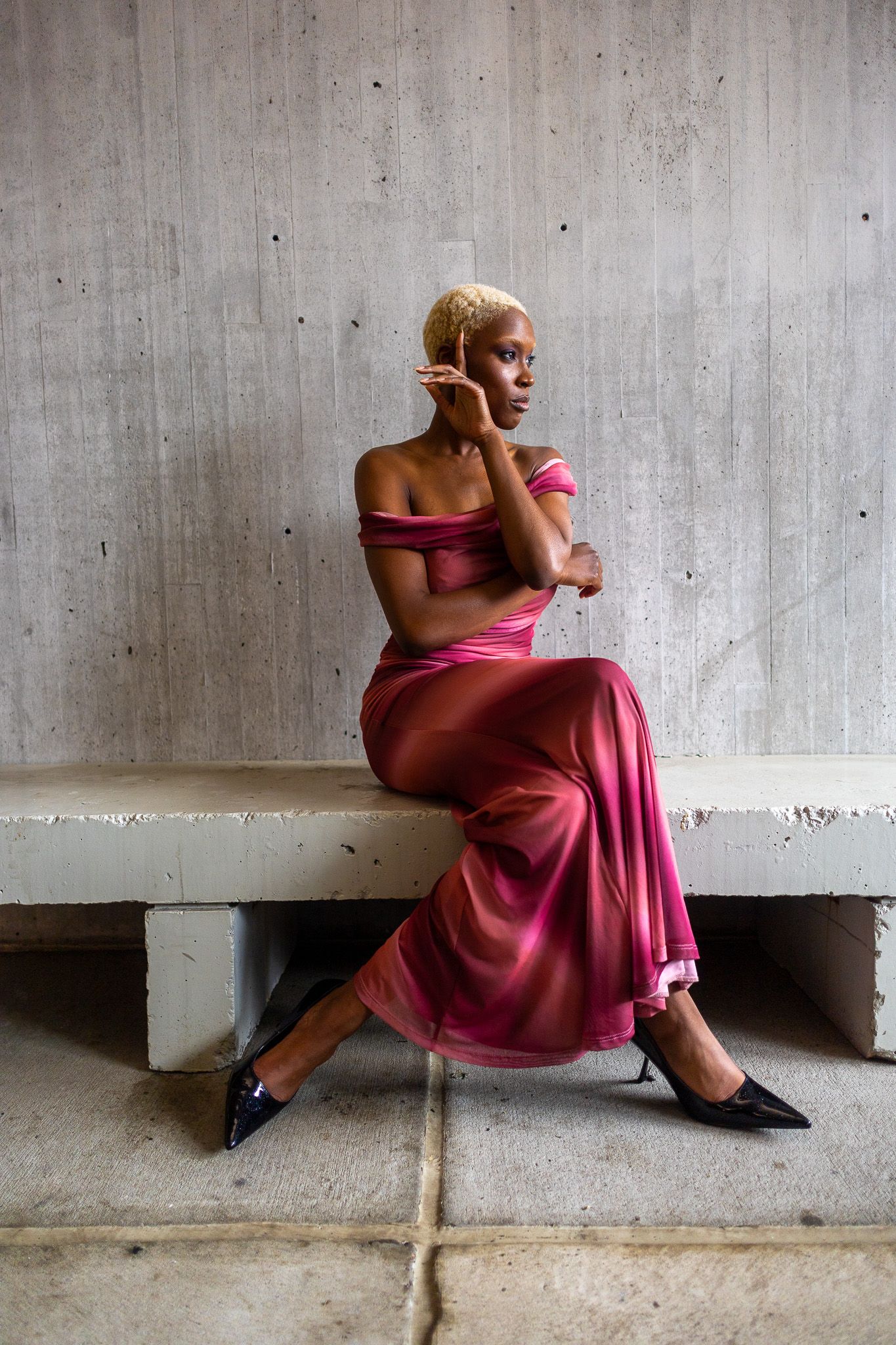 A woman in a strapless, flowing magenta dress and black heels is sitting on a concrete bench against a textured gray wall.