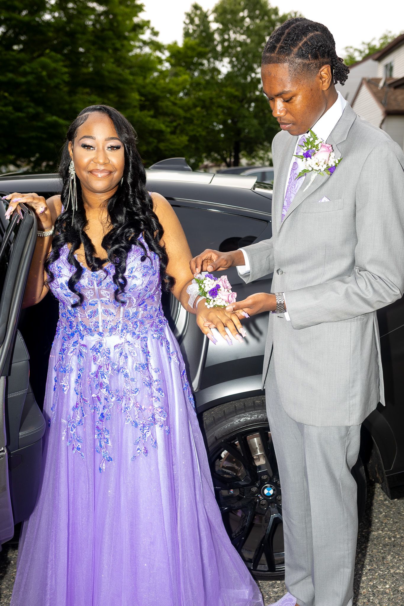 This image features a woman in a purple dress standing next to a man in a gray suit, both positioned near a dark vehicle.