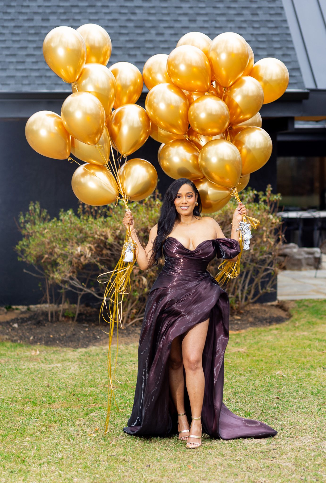 woman standing outdoors, holding a large bunch of gold balloons. She is wearing a dark, elegant dress with a high slit, and on grass.