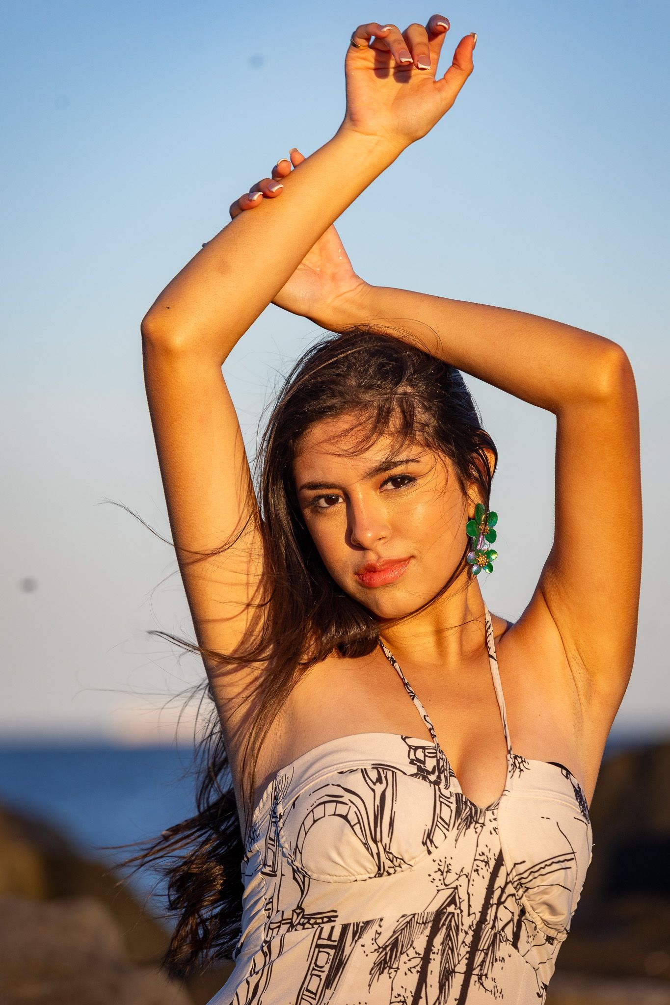 a woman standing outdoors, posing with her arms raised above her head. She is wearing a strapless, patterned top and large hoop earrings.