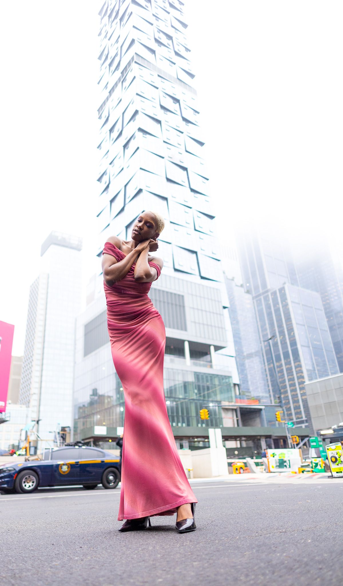 a woman in a long, flowing pink dress standing on a city street, with a tall, modern skyscraper rising in the background.