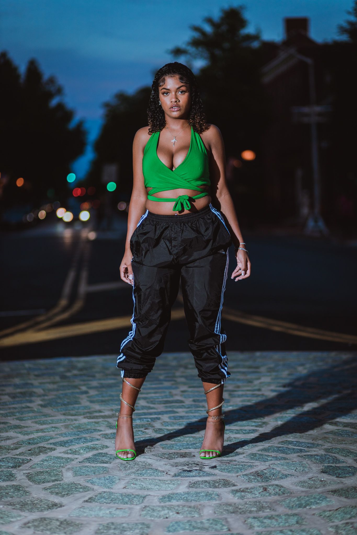 a woman standing outdoors at night, wearing a green top and dark pants. She is illuminated against a dark background with a building.