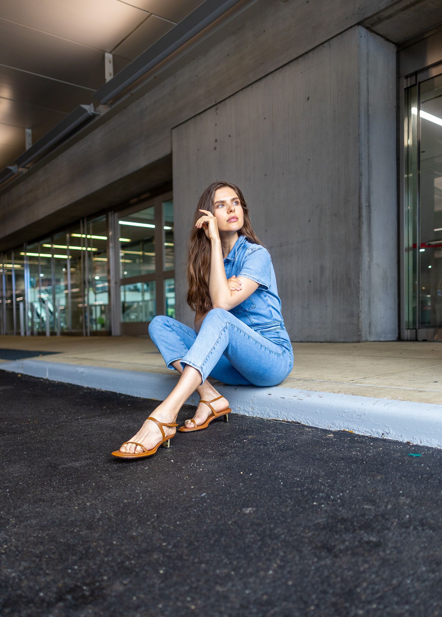 A woman in a blue denim jumpsuit and brown sandals is sitting on a ledge in front of a modern gray building.