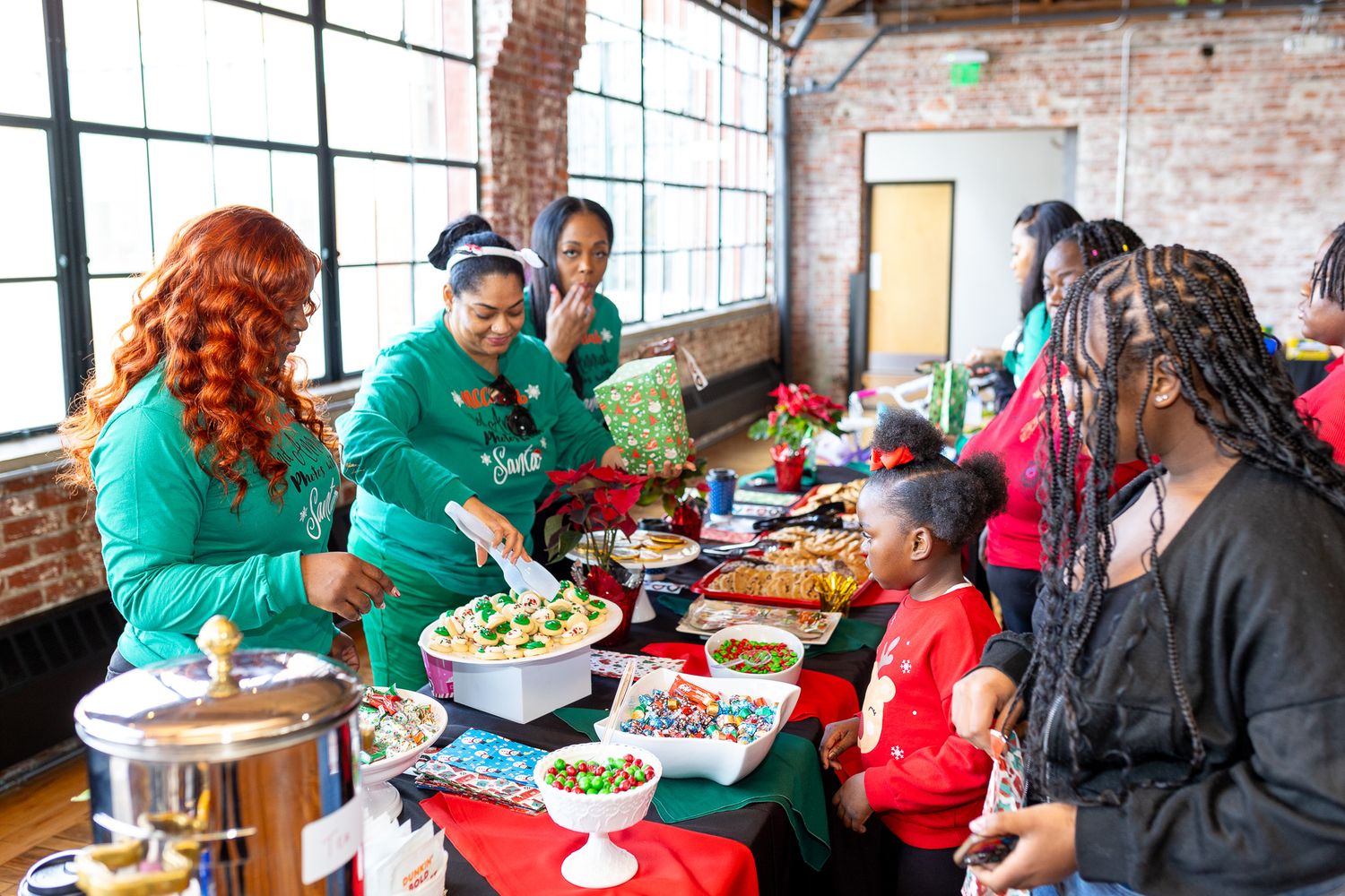 This photo features a group of people gathered around a table filled with food, with two women in green shirts appearing to serve.