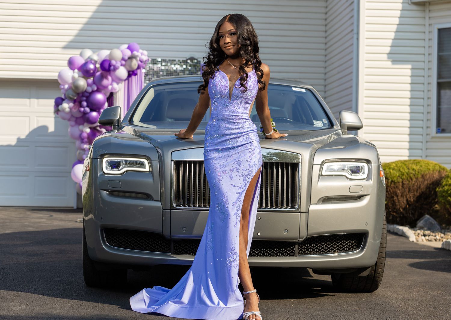 This image shows a woman in a long, flowing light blue dress with a high slit, posing in front of a silver Rolls-Royce. There are purple.