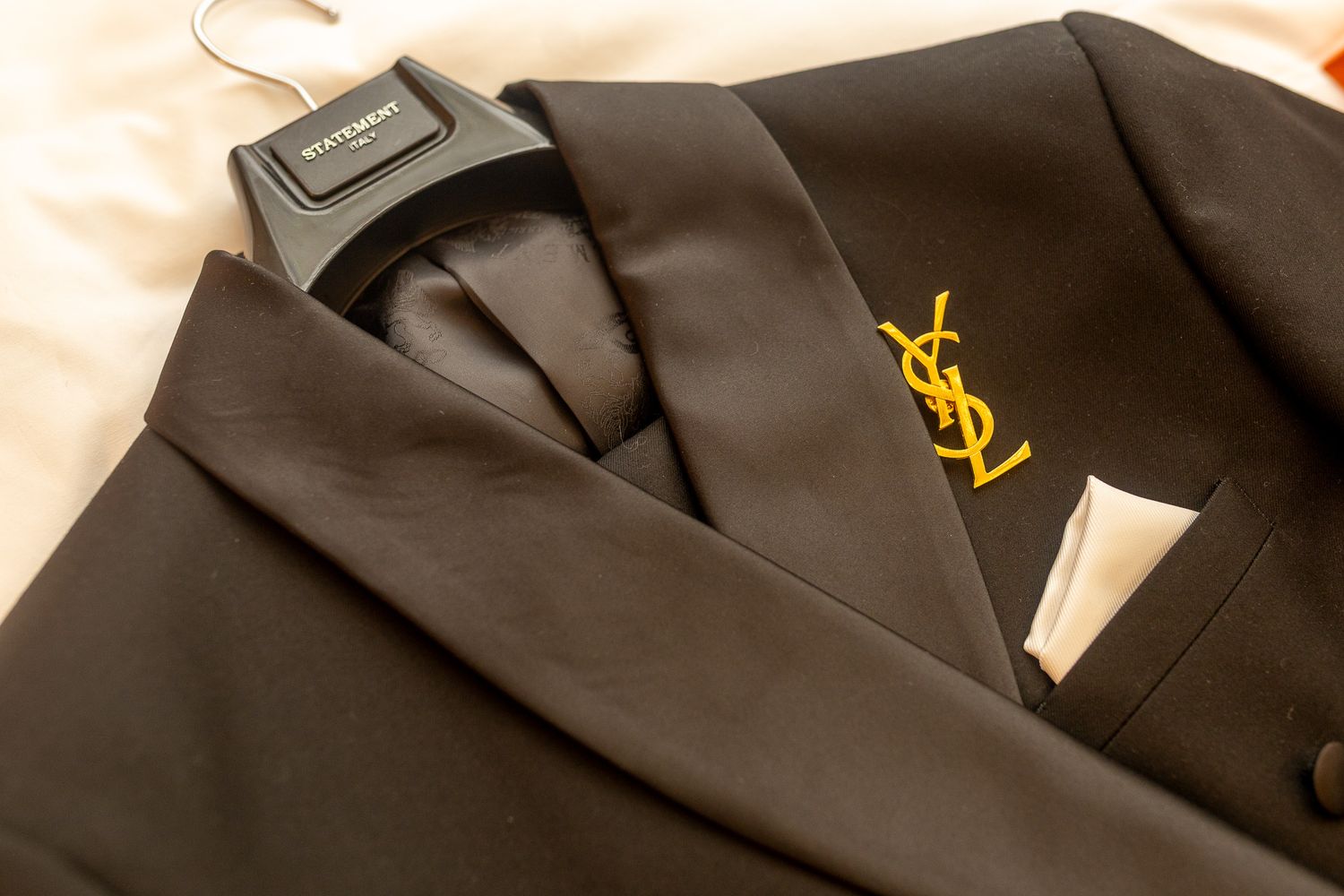 This image features a close-up of a black Saint Laurent suit jacket, highlighting the gold YSL logo on the lapel and a white pocket square.