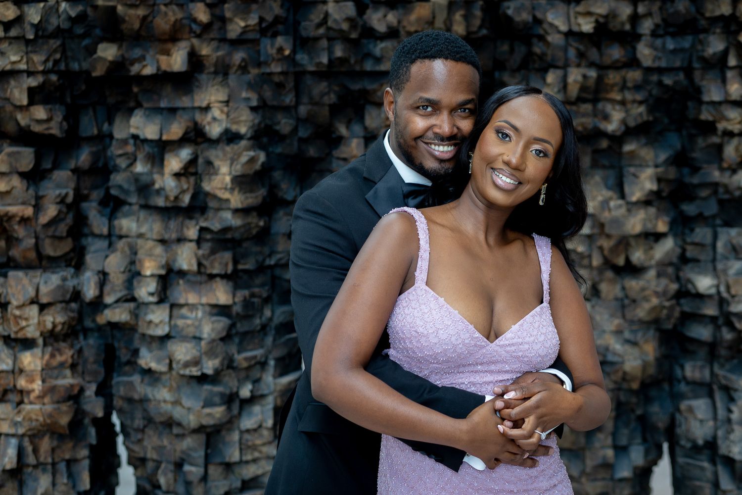 a man and a woman posing together against a dark, textured background. Engagement photoshoot with  dark suit, and a light purple dress.