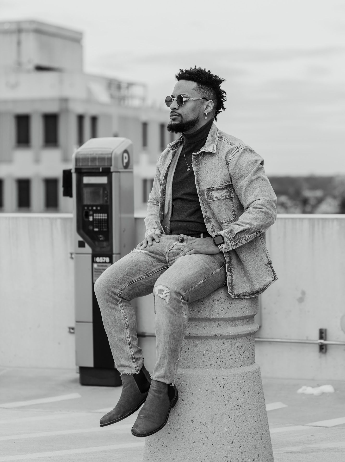 This is a black and white photo of a man sitting on a ledge outdoors, wearing a jacket, jeans, and sunglasses, with a building in the backgr