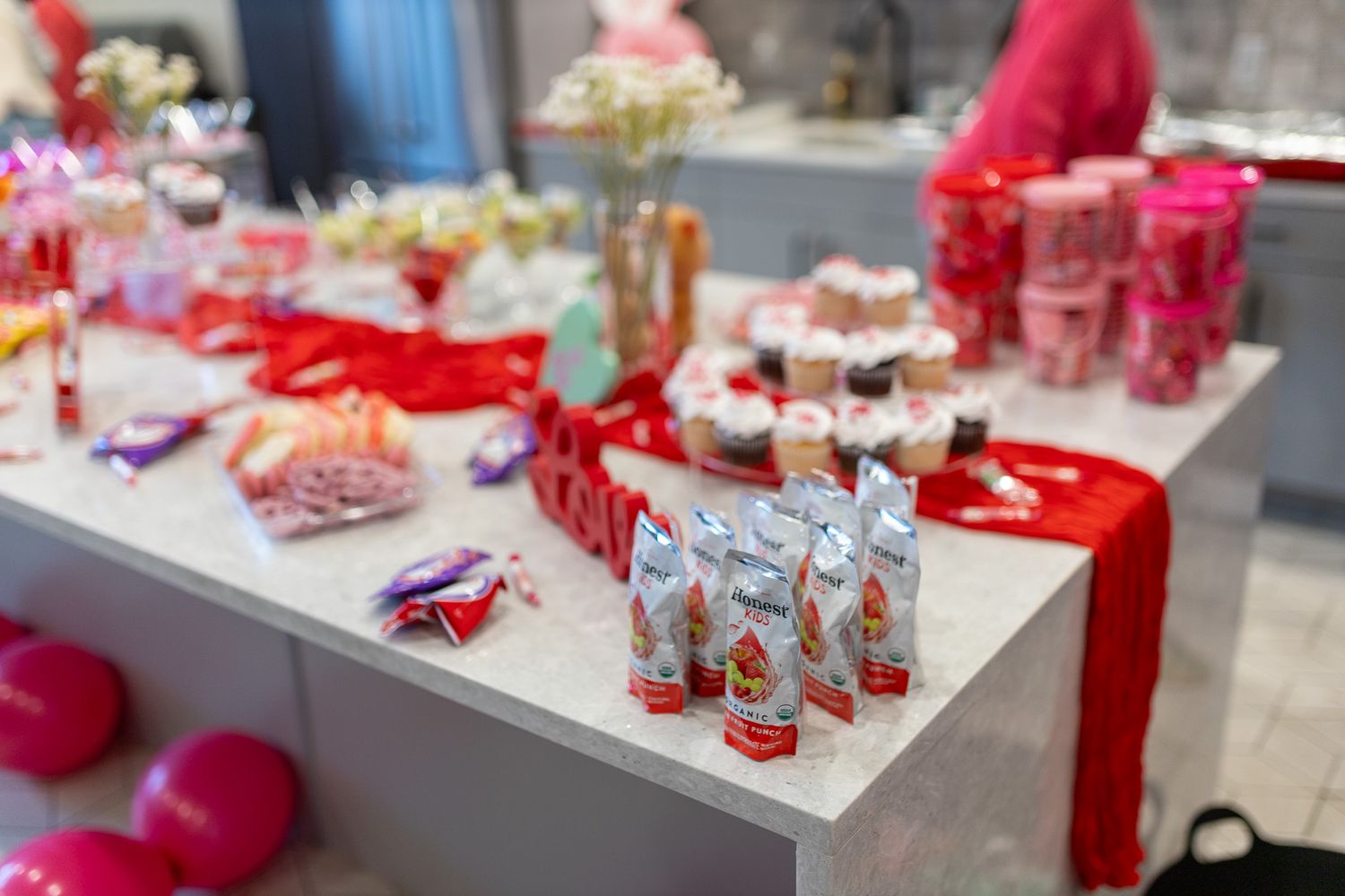 This image shows a dessert table set up for an event, featuring a red table runner and various treats including cupcakes, cake pops.