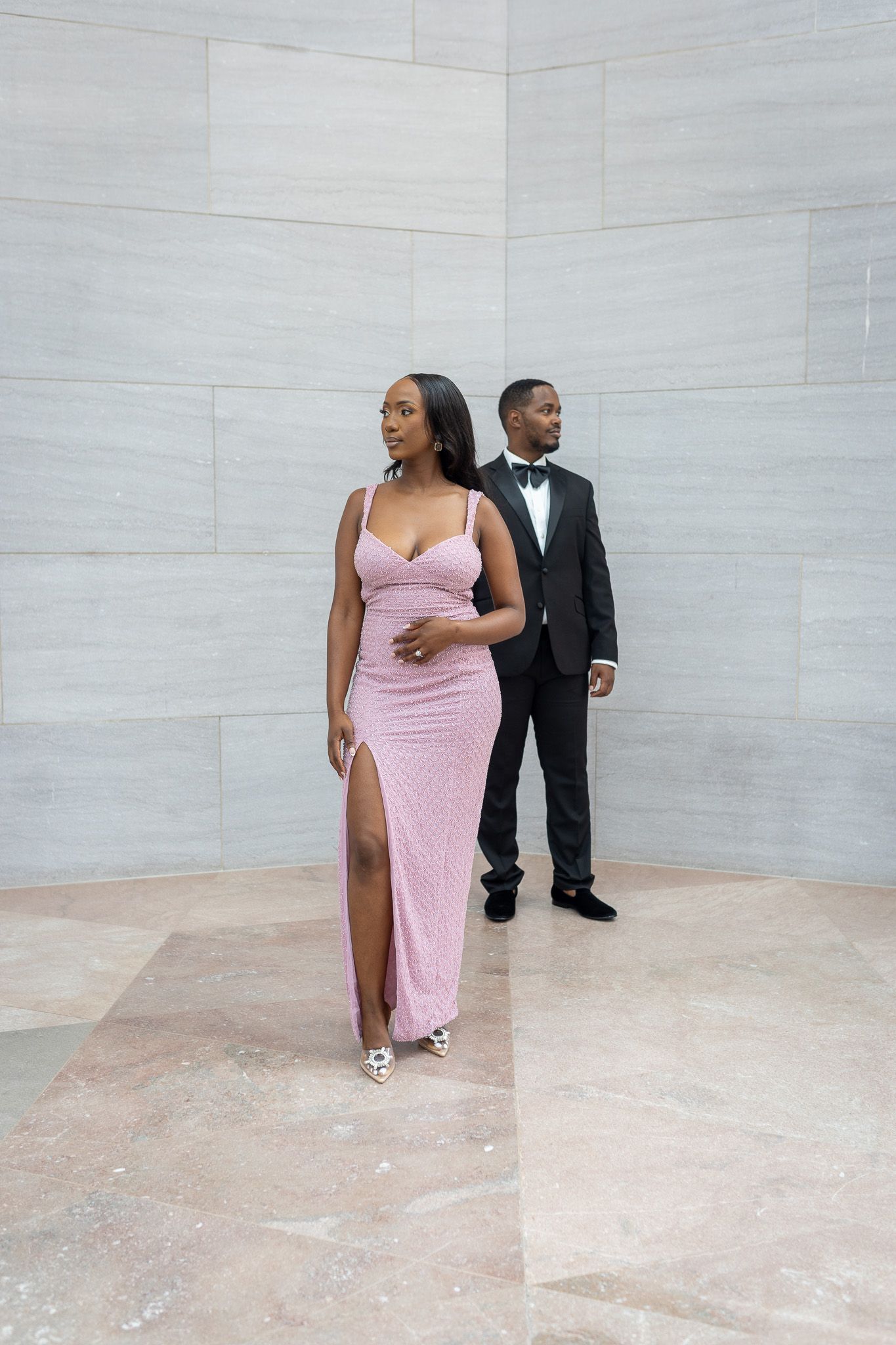 a woman in a long, light pink dress with a high slit, standing next to a man in a black tuxedo. Engagement photoshoot at an art museum.