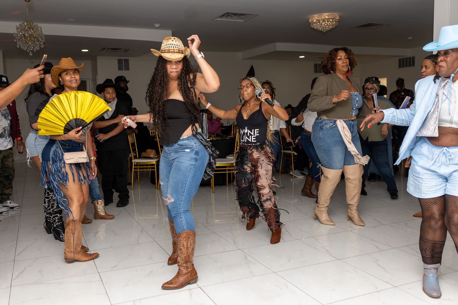 a group of people dancing indoors. They appear to be line dancing or following a routine, with several individuals wearing cowboy hats.
