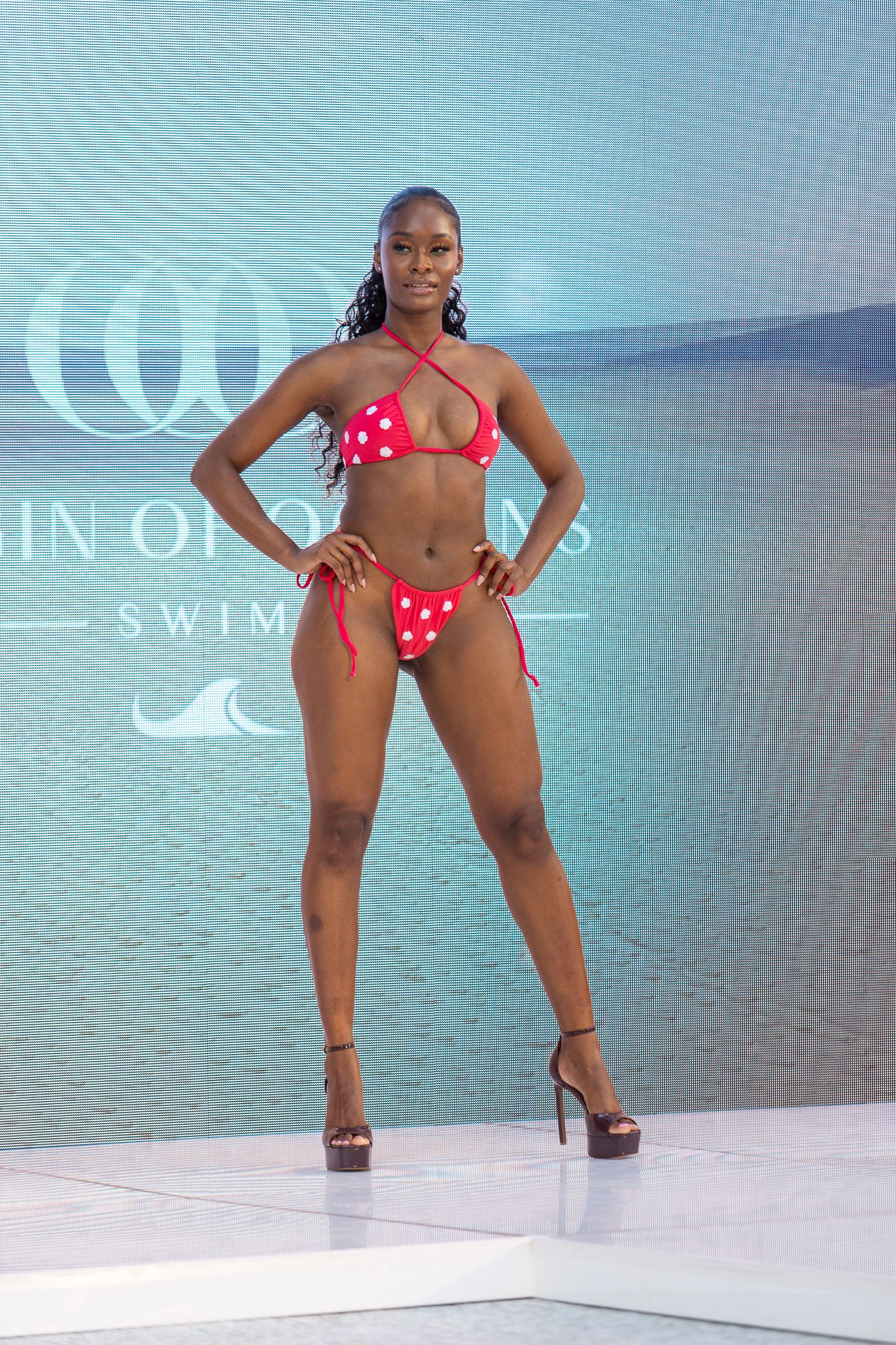 This photo features a model walking down a runway wearing a red and orange patterned bikini.