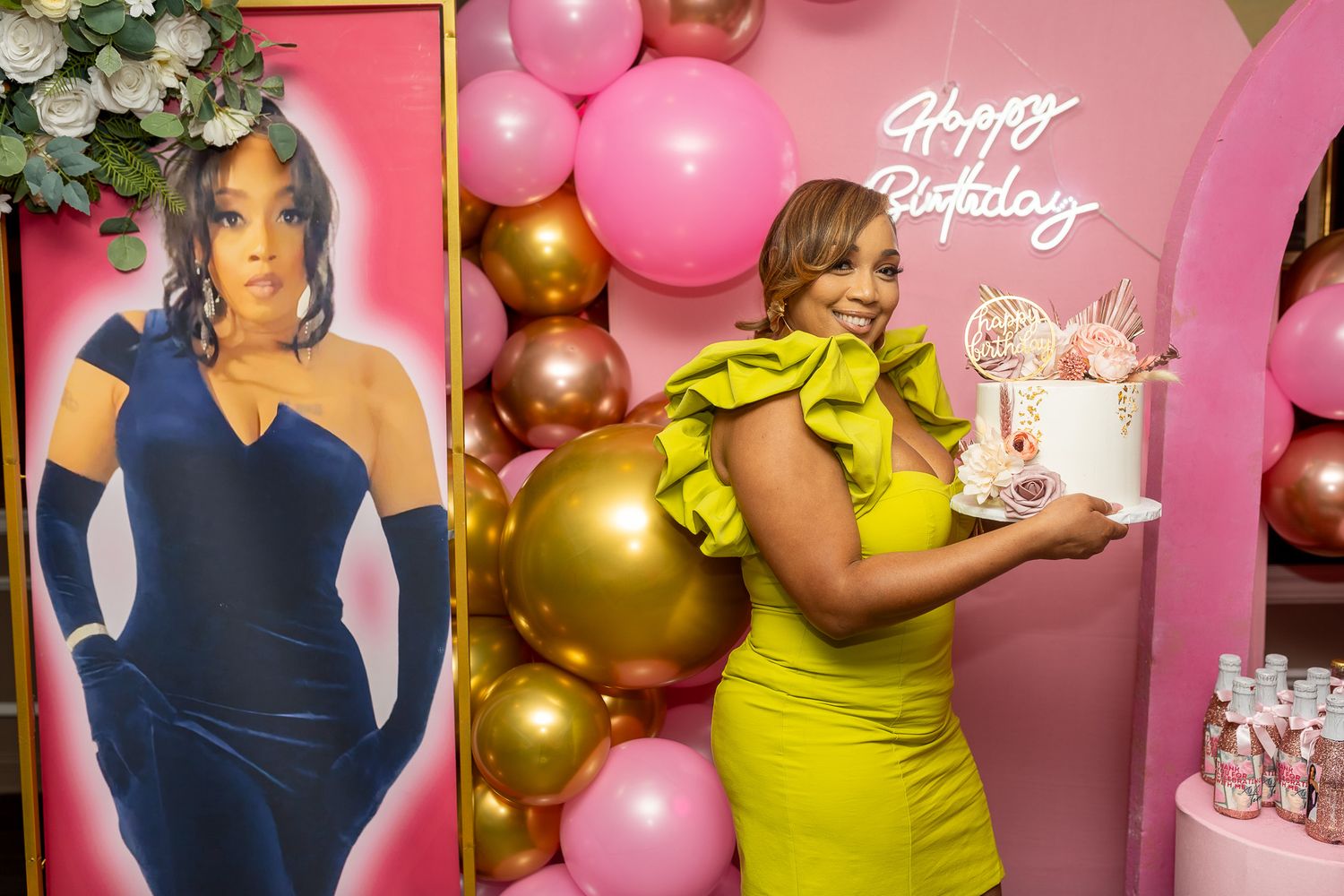 a woman in a bright yellow dress holding a birthday cake, standing next to a life-sized cutout of a woman in a blue dress.