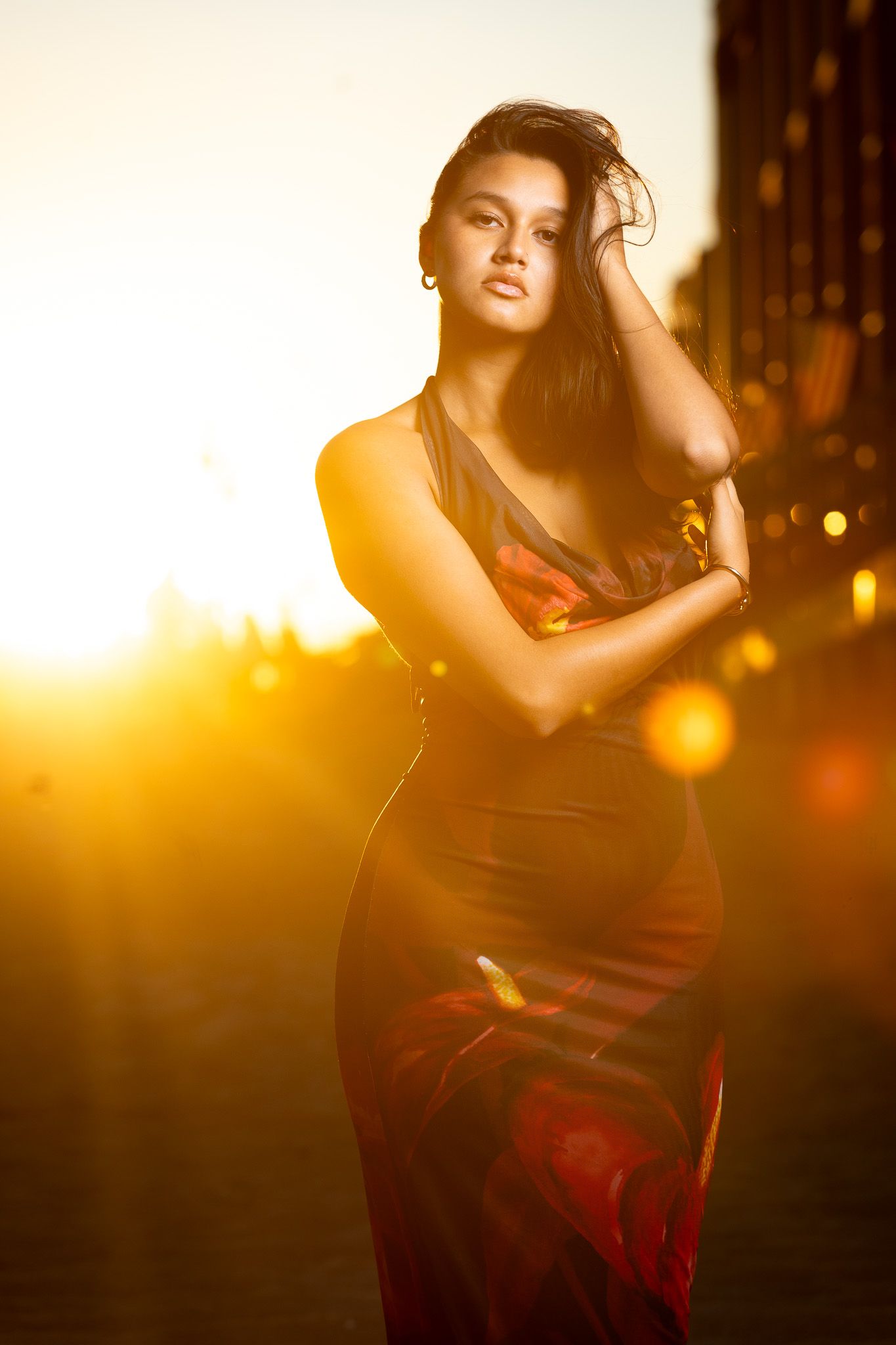 a woman in a long, flowing red dress standing on a beach at sunset. The warm, golden light of the setting sun creates a backlit effect.