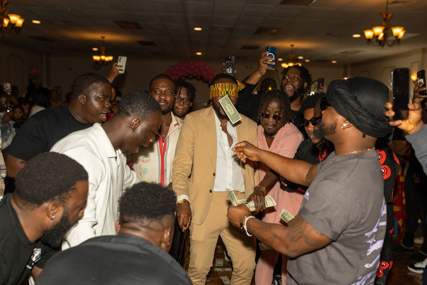 This photo features a group of people gathered indoors, likely at a social event or celebration. A man in a tan suit stands in the center.