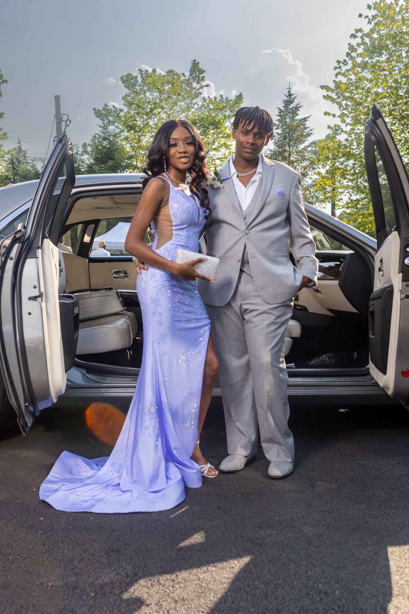 This image shows a couple posing next to a white car. The woman is wearing a long, flowing lavender dress, and he is wearing a light suit