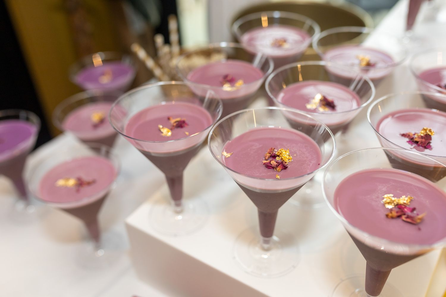 several martini glasses filled with a vibrant purple beverage, garnished with small edible flowers or herbs, arranged on a white tray.