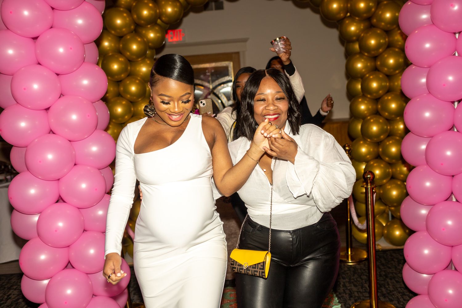Two women posing together in front of a backdrop of pink and gold balloons. One woman is wearing a white long-sleeved dress. A baby shower.