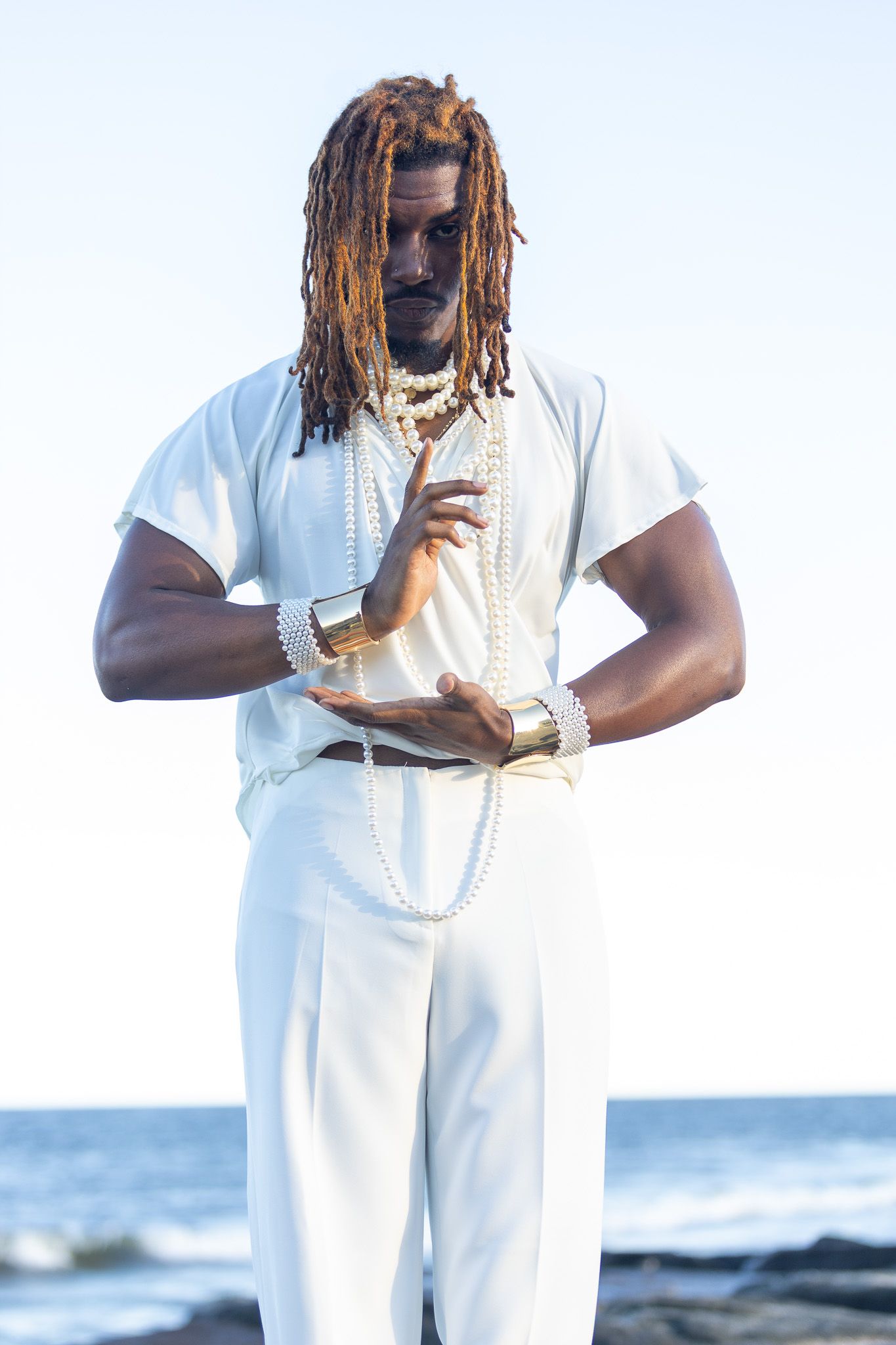 a man with long dreadlocks standing on  beach stones with the ocean in the background. He is wearing an all-white outfit  and posing hands.