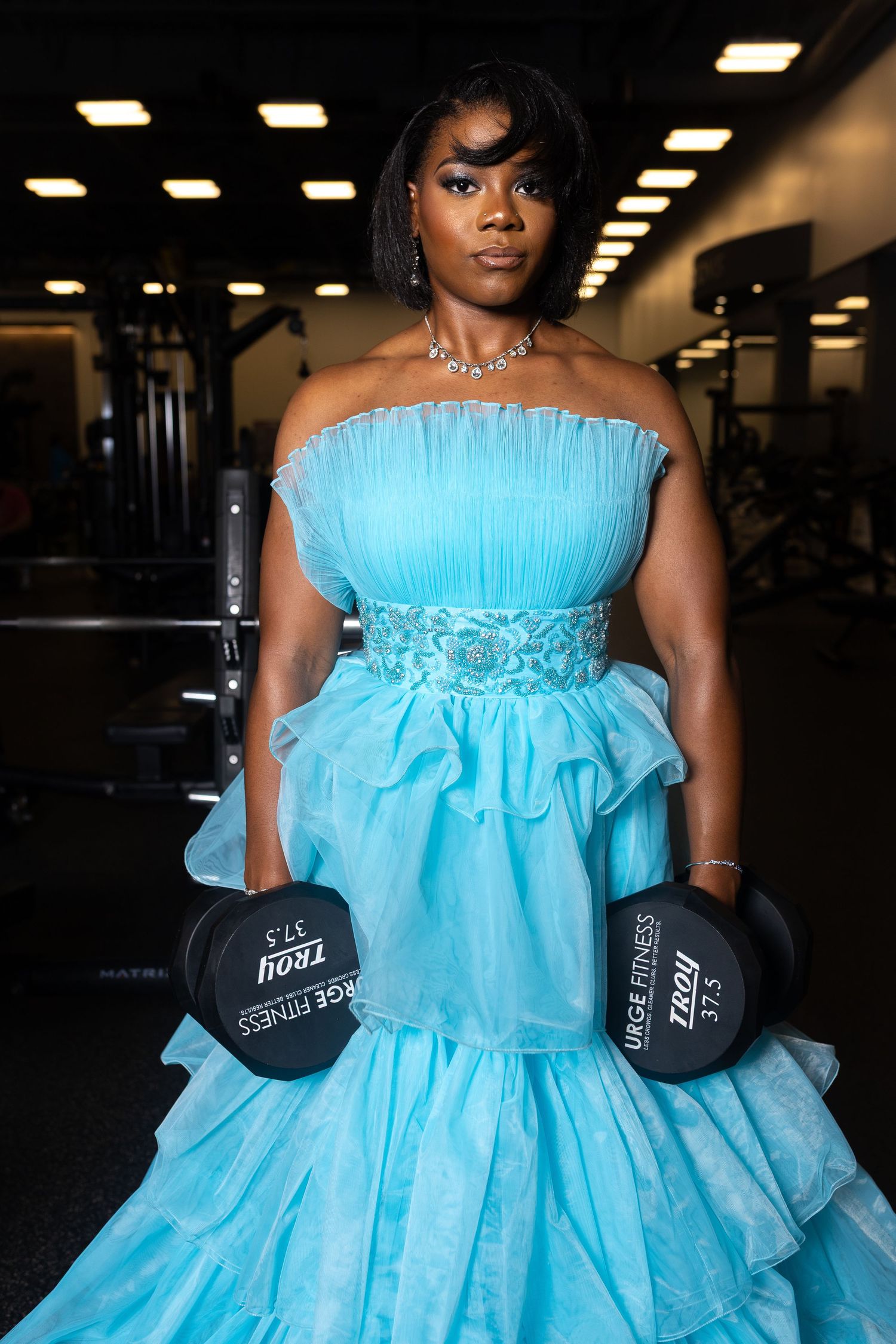a woman standing with her back to the camera, wearing a strapless, tiered, light blue tulle prom grown gown while lifting weights in a gym.