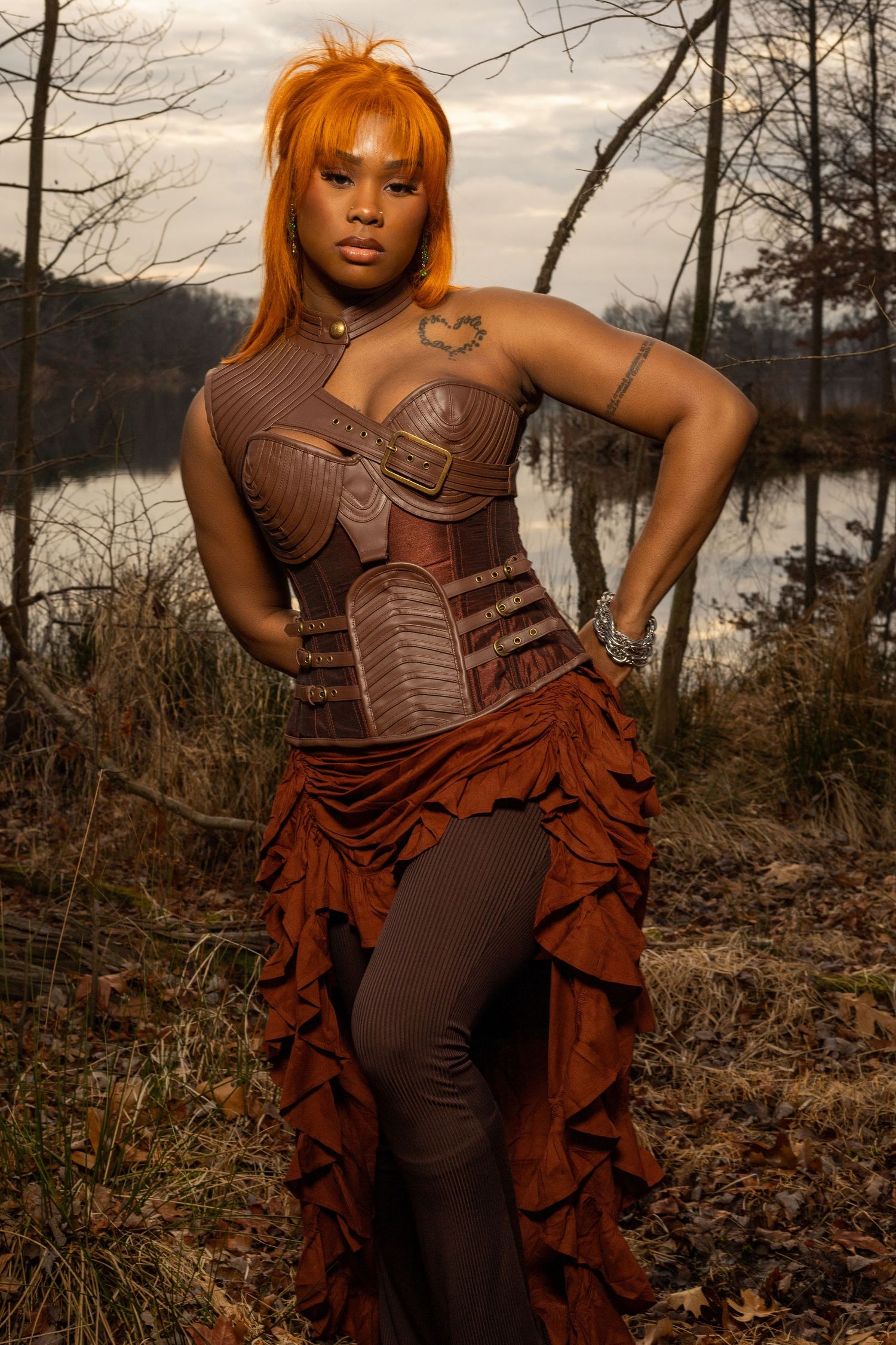 This photo features a woman with vibrant orange hair posing outdoors near a body of water and bare trees. She is wearing a brown outfit.