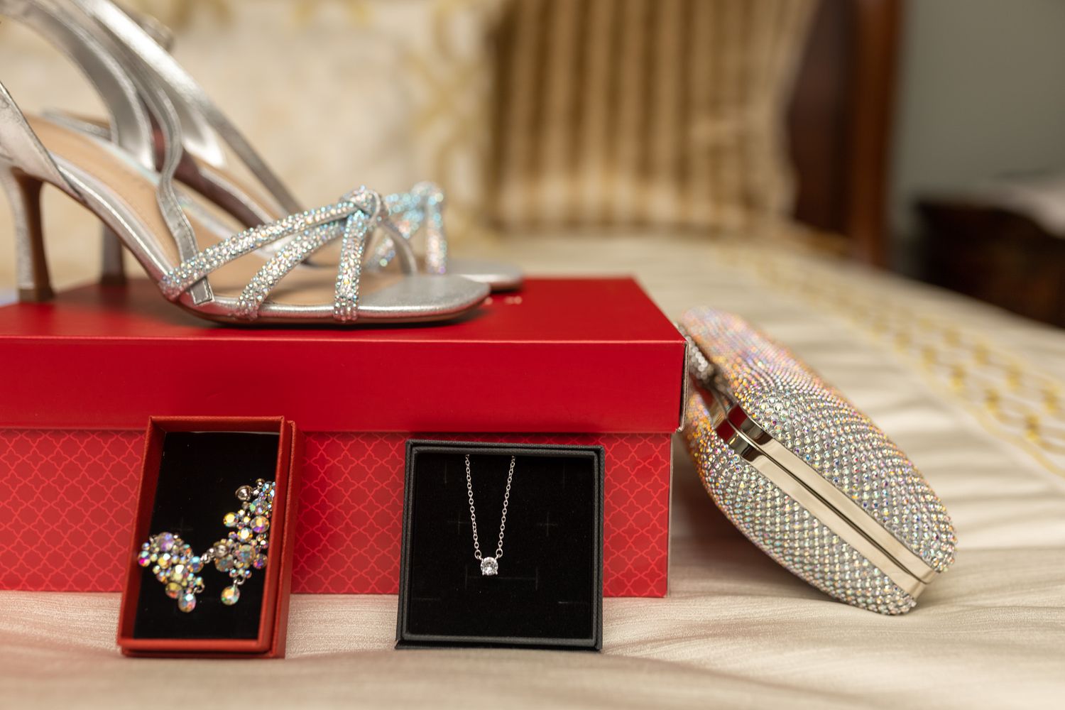 This image features a pair of silver, strappy high-heeled shoes resting on a red box, accompanied by two small jewelry boxes,