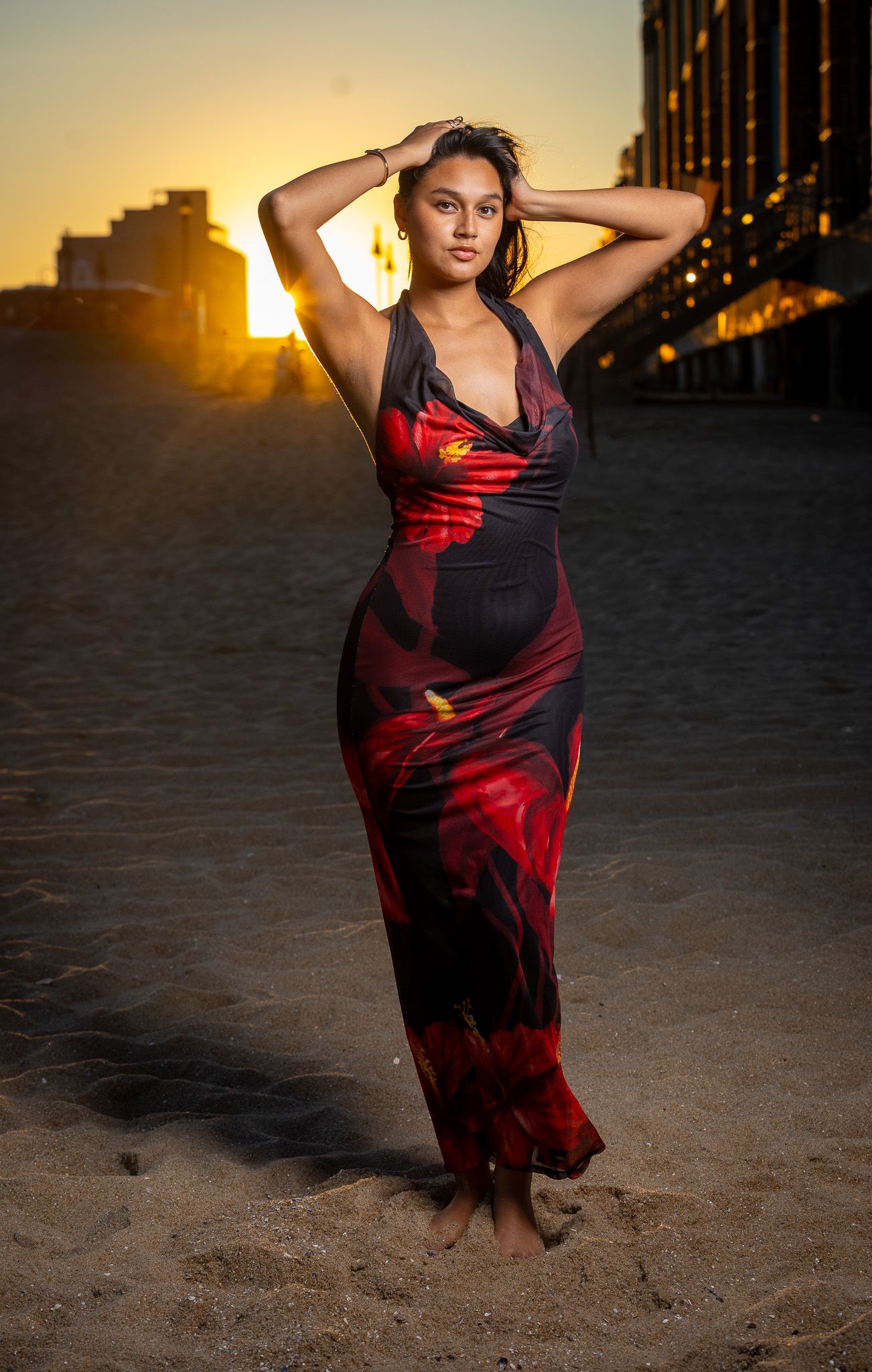 A woman in a long, flowing black and red dress stands on a beach at sunset, with her hands raised to her head. Used off camera flash.
