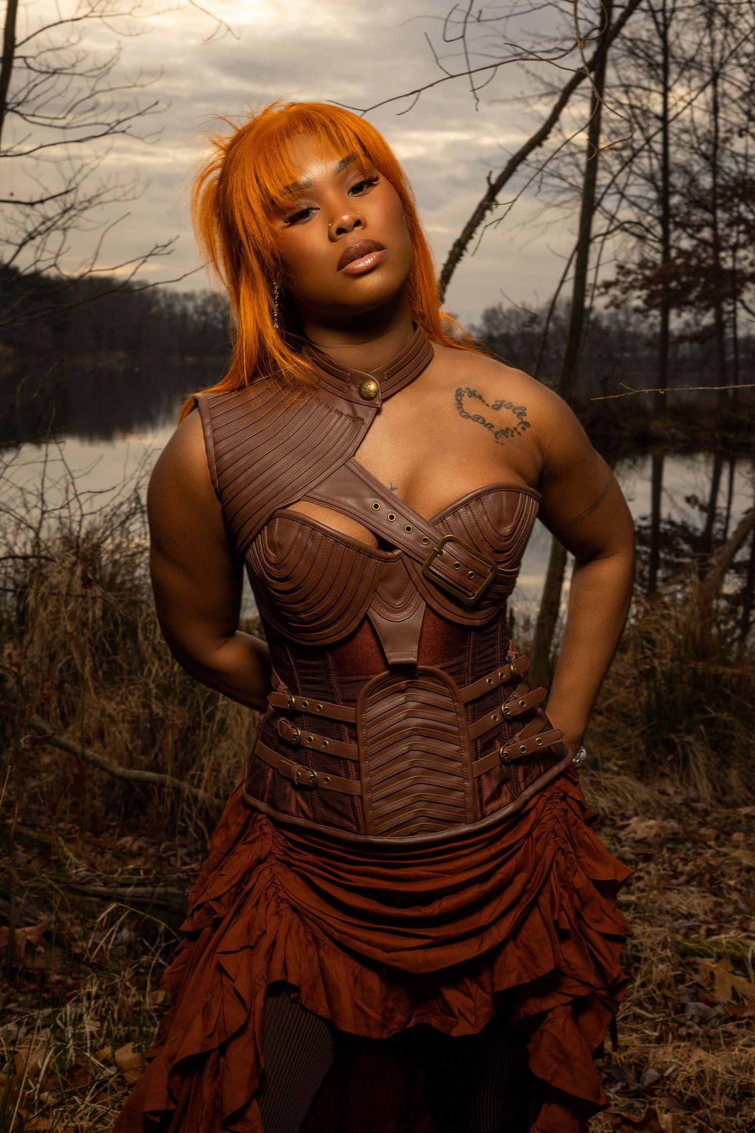 woman with long, vibrant orange hair standing outdoors in a wooded area. Wearing a dark, textured, strapless dress with a background lake.