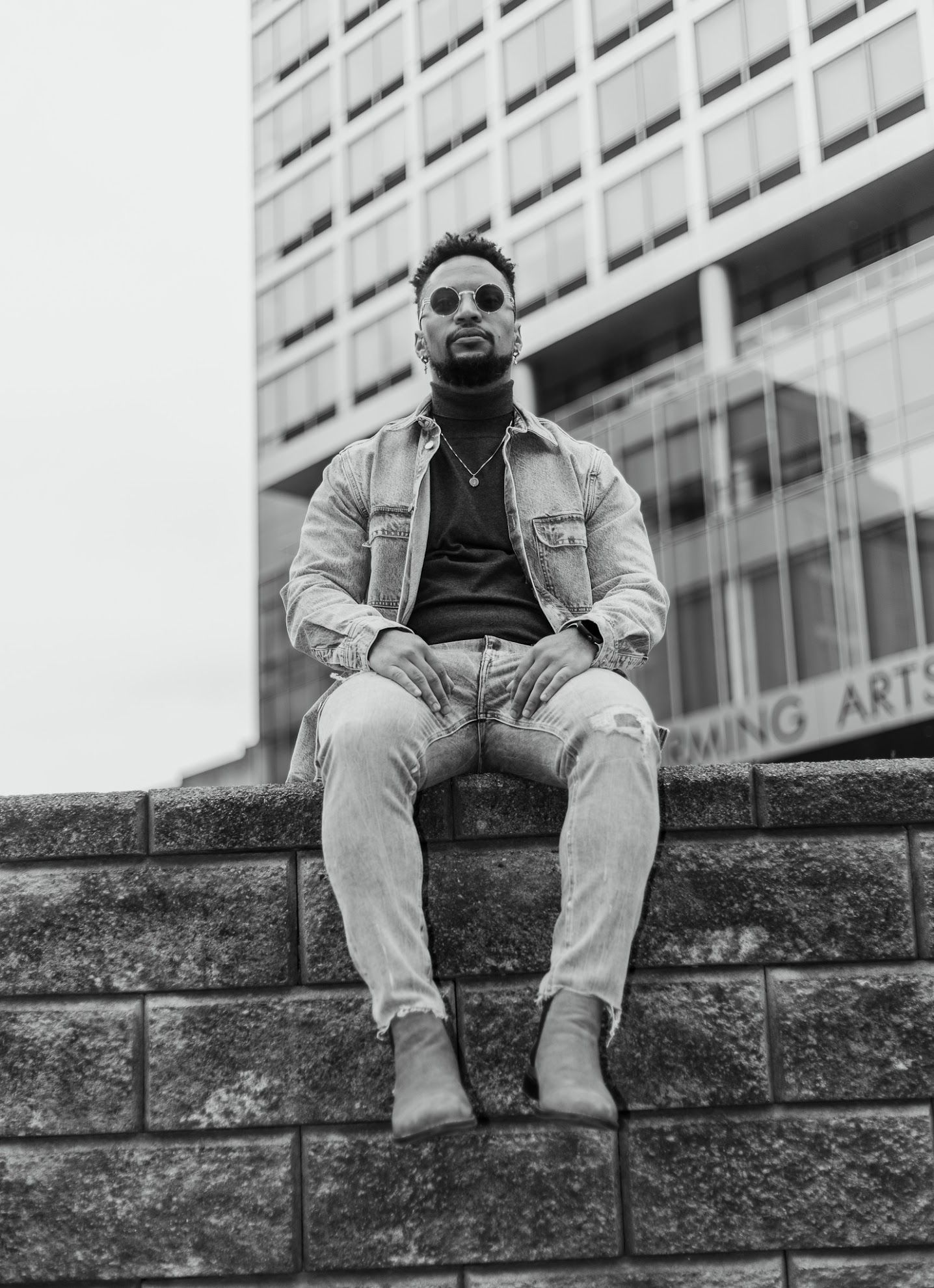 This black and white photo features a man sitting on a ledge in front of a building, wearing a jacket, jeans, and sunglasses.