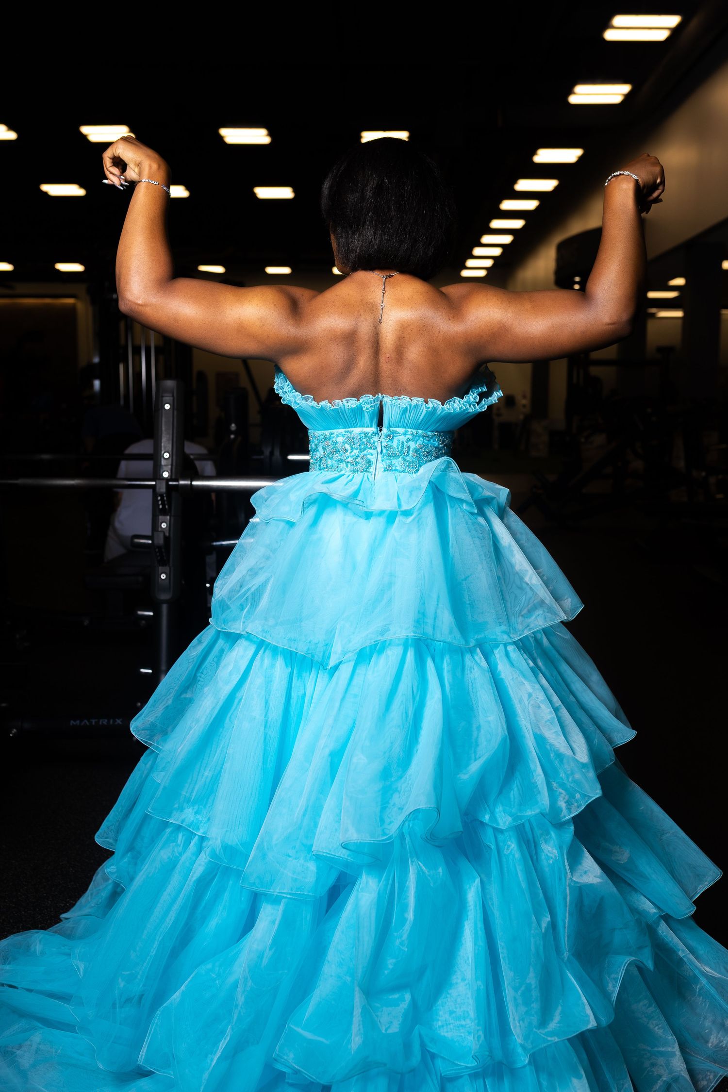 a woman standing with her back to the camera, wearing a strapless, tiered, light blue tulle prom gown while flexing her muscles in a gym.