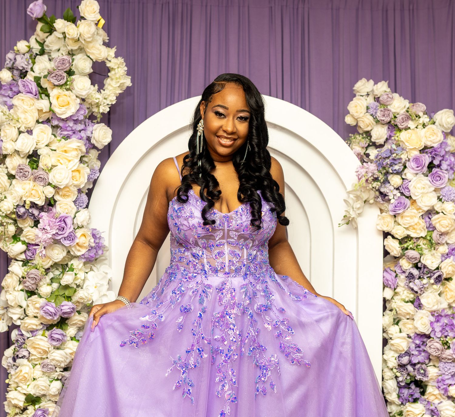 This image features a woman in a strapless, flowing purple gown, standing in front of a white arch adorned with white and purple flowers.