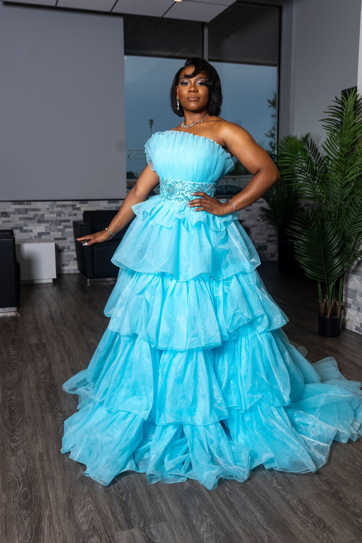 A woman wearing a strapless, tiered, light blue tulle prom gown stands indoors at a gym, posing with one hand on her hip. A prom sendoff.