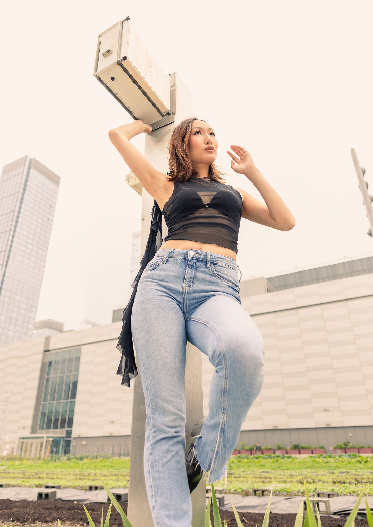 A woman in a black top and light blue jeans stands outdoors, looking up, with a modern building in the background.