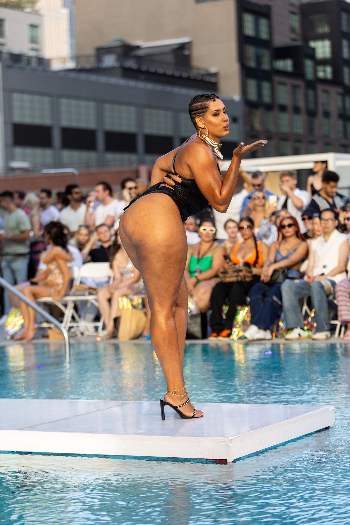 This photo features a woman standing by a swimming pool, wearing a black bikini top and bottoms with black high heels at NY Swim Week.