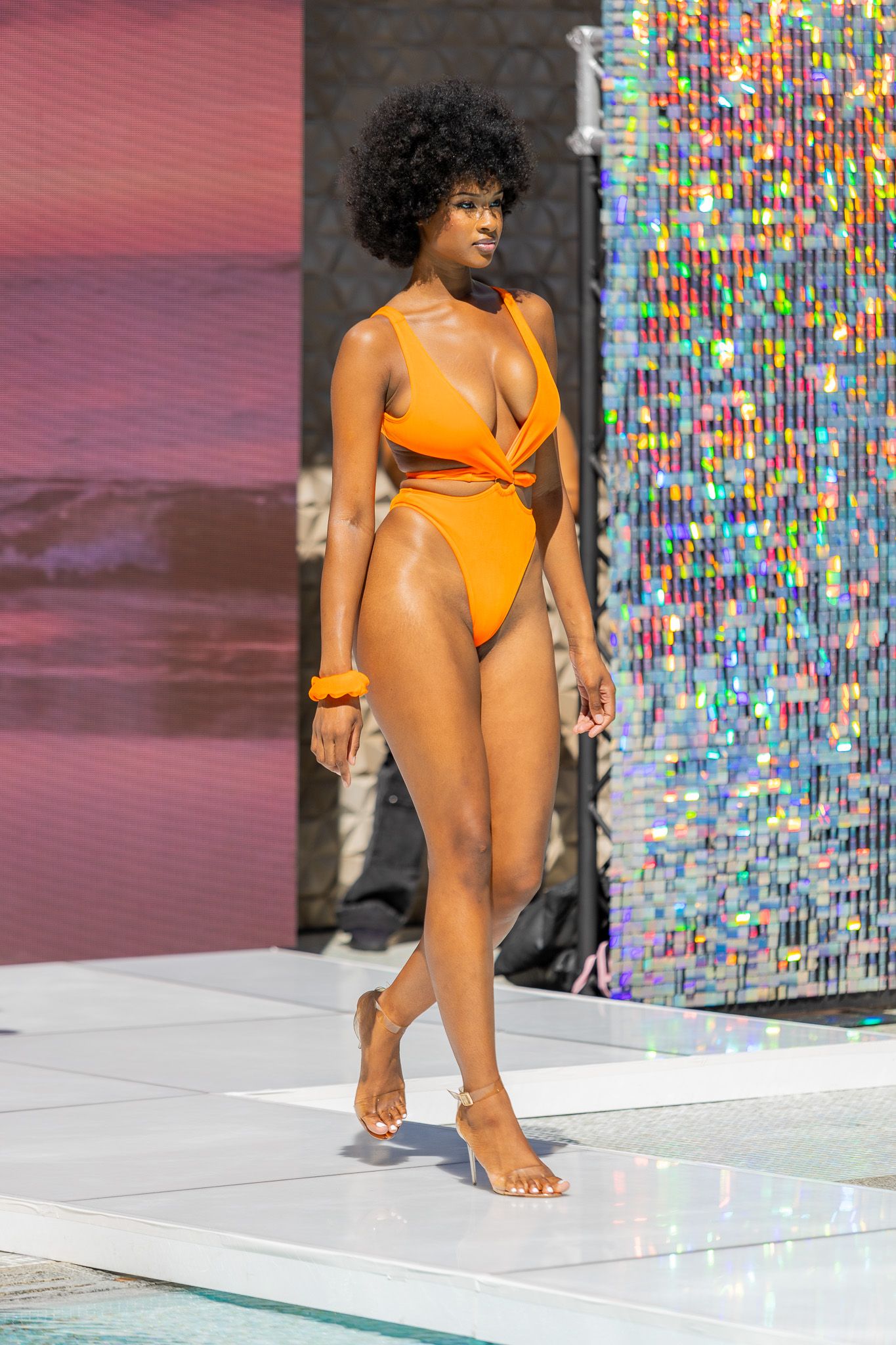 This photo features a model walking down a runway wearing an orange bikini.