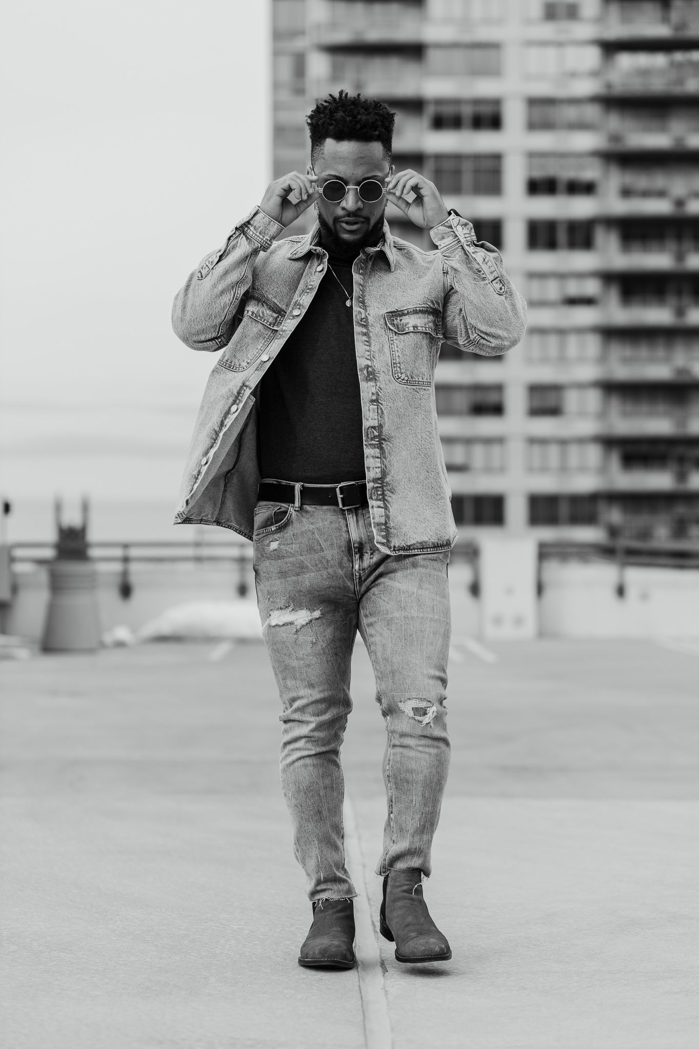 This black and white photo features a man standing outdoors, wearing a jacket, jeans, and sunglasses, with a building in the background.