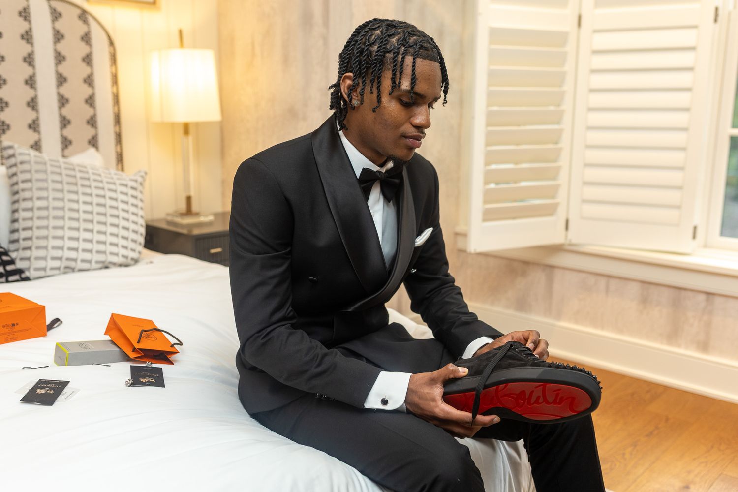 This image features a man in a black suit sitting on a bed, holding a black dress shoe with a red sole.