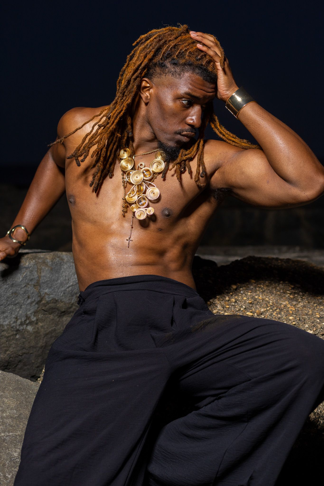 a shirtless man with long dreadlocks, wearing dark pants and a gold necklace, posing against a dark background with his hand raised.